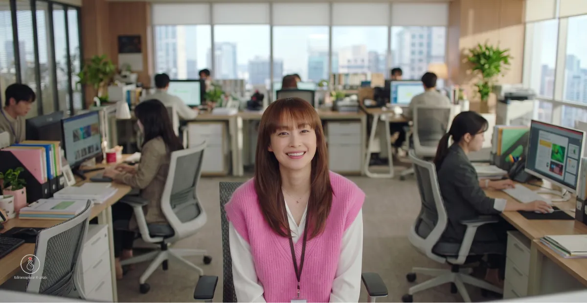 Cena panorâmica do dorama Romance is a Bonus Book com a atriz Lee Na-young vestindo colete rosa e sorrindo no escritório da editora Gyeorye.