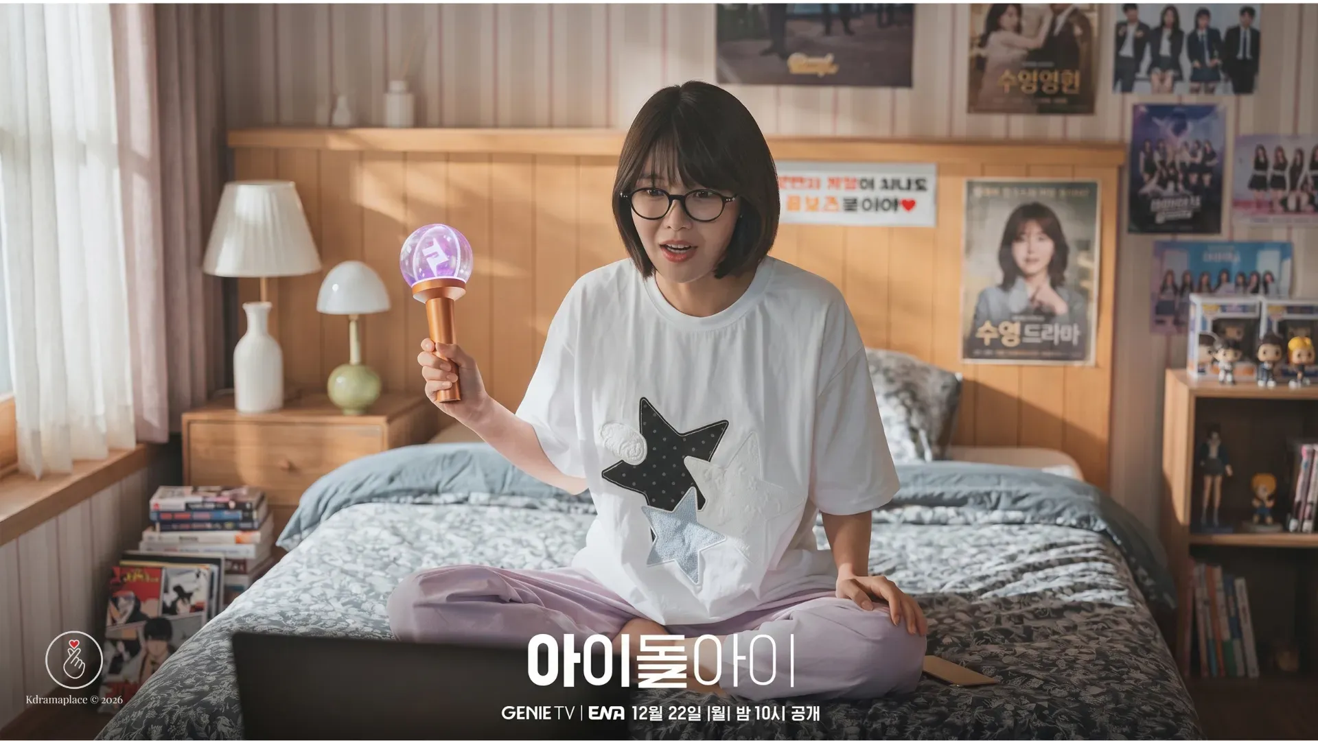 Cena do dorama Meu Ídolo onde a atriz Choi Soo-young interpreta uma fã de óculos sentada na cama de seu quarto decorado com pôsteres, segurando um lightstick roxo iluminado com expressão de empolgação enquanto assiste a uma transmissão no notebook.