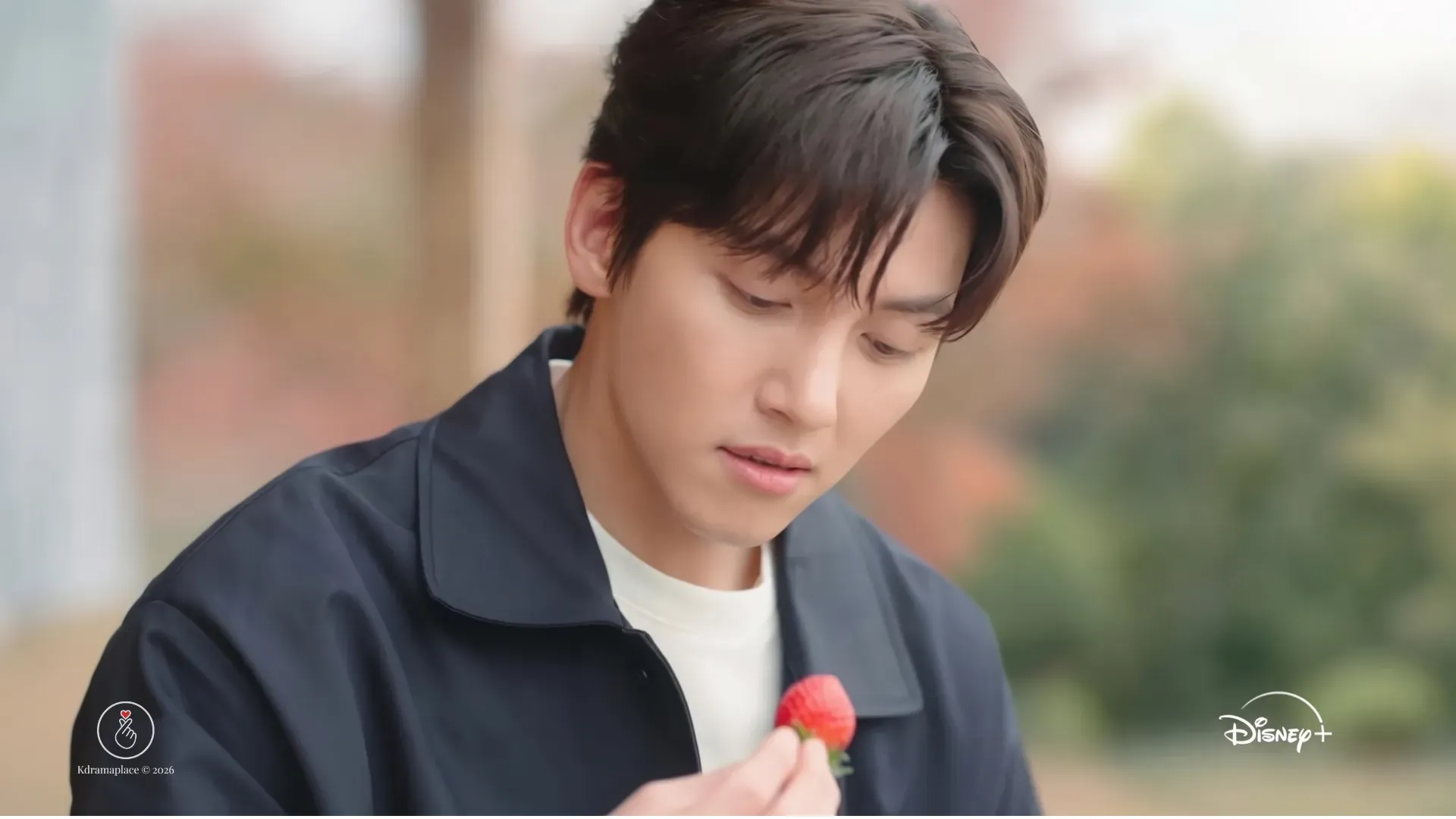 Close-up do ator Ji Chang-wook no dorama Merry Berry Love, vestindo um casaco preto e camiseta branca, olhando pensativo para um morango fresco que está segurando em sua mão, com o logotipo da Disney+ no canto inferior direito.
