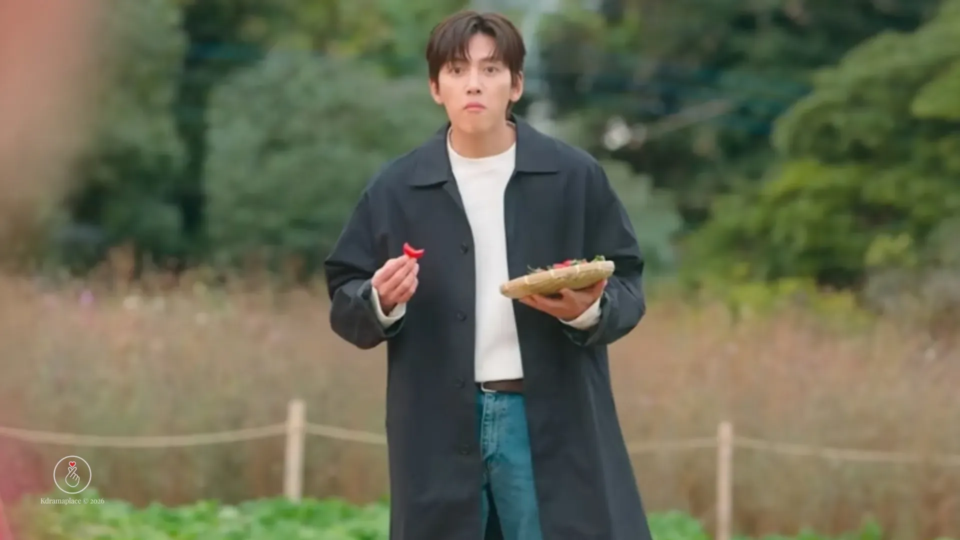 Cena de Merry Berry Love mostrando o ator Ji Chang-wook em pé no meio de um campo aberto, vestindo um sobretudo preto sobre calça jeans, segurando uma pequena cesta trançada de morangos enquanto olha para frente com uma expressão curiosa.