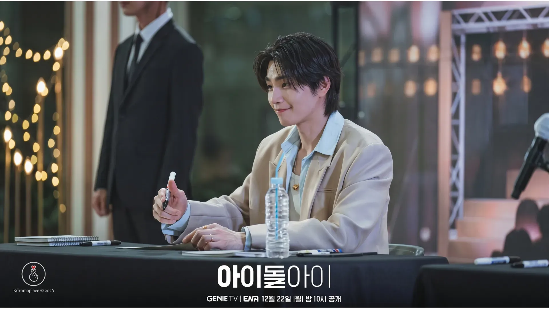  Cena de Meu Ídolo mostrando o ator Kim Jae-young com um sorriso charmoso, vestindo um blazer bege claro em uma mesa de evento de autógrafos com fãs, segurando uma caneta com garrafa de água à frente e um segurança de terno ao fundo.