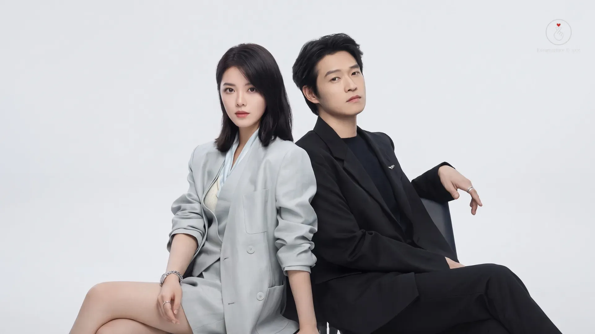 Atriz Zhao Jinmai de terno cinza e o ator Wei Daxun de terno preto sentados de costas um para o outro em fundo branco.